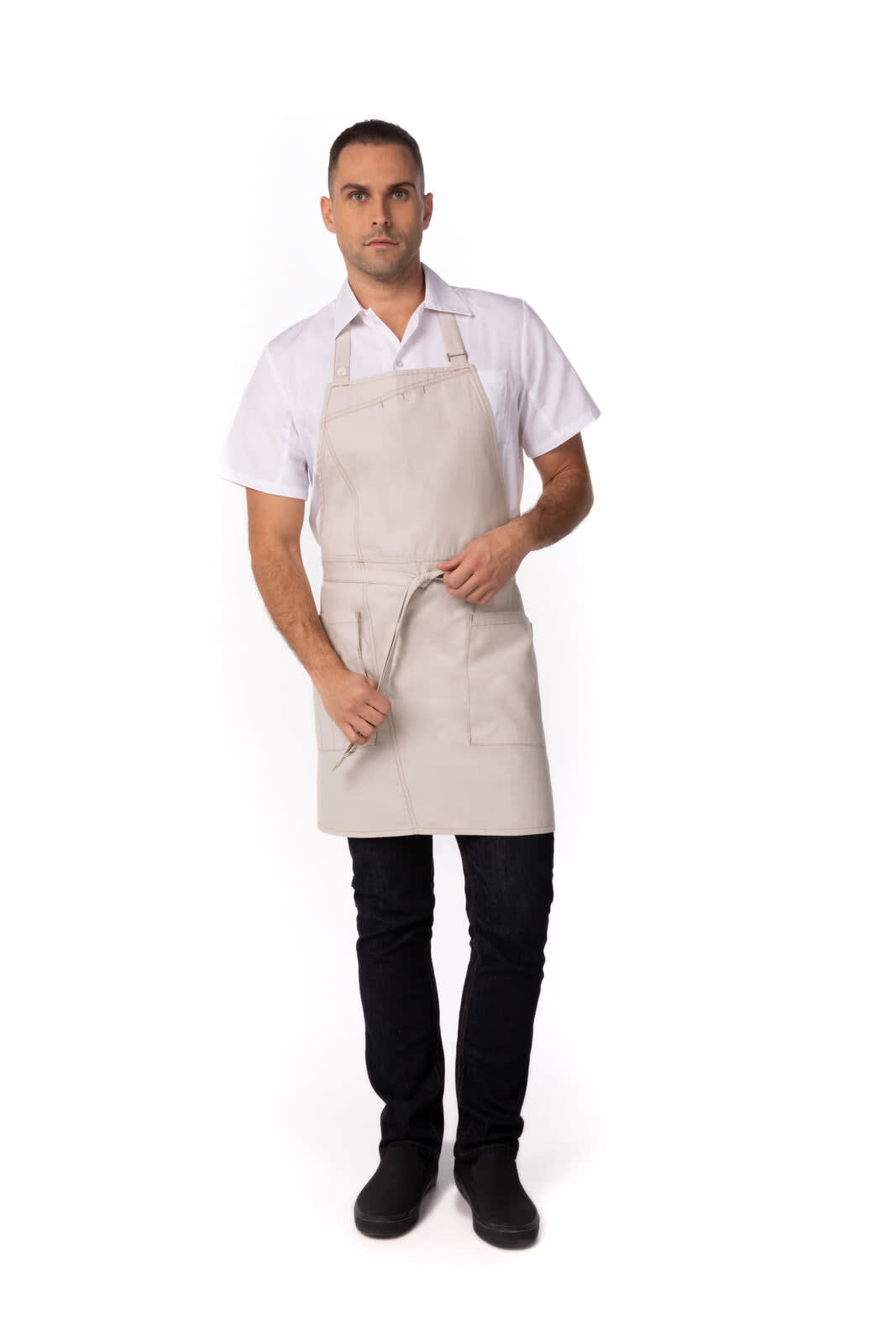 A man wearing a light grey Medford Bib Apron, featuring a logo and a belt tie, stands against a plain background.