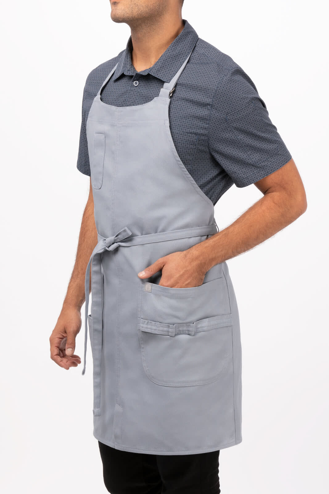 A grey bib apron with pockets, worn by a person standing sideways, featuring an adjustable neck strap.