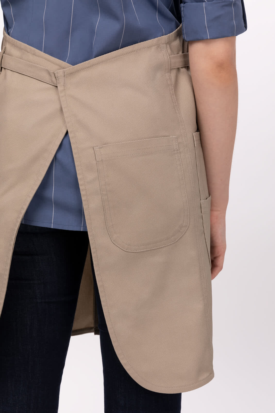 A khaki Logan Bib Apron with pockets, worn over a blue striped shirt and jeans. The apron features a cross-back design.