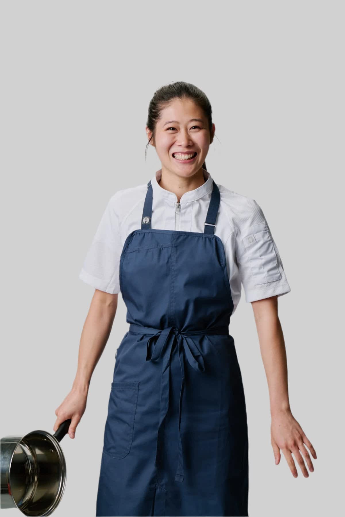 A smiling individual wearing a navy blue Brio Bib Apron, holding a pot, with a logo on the apron.