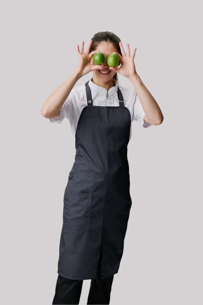 A dark grey bib apron worn by a person holding two green apples, smiling playfully. The apron features adjustable straps.