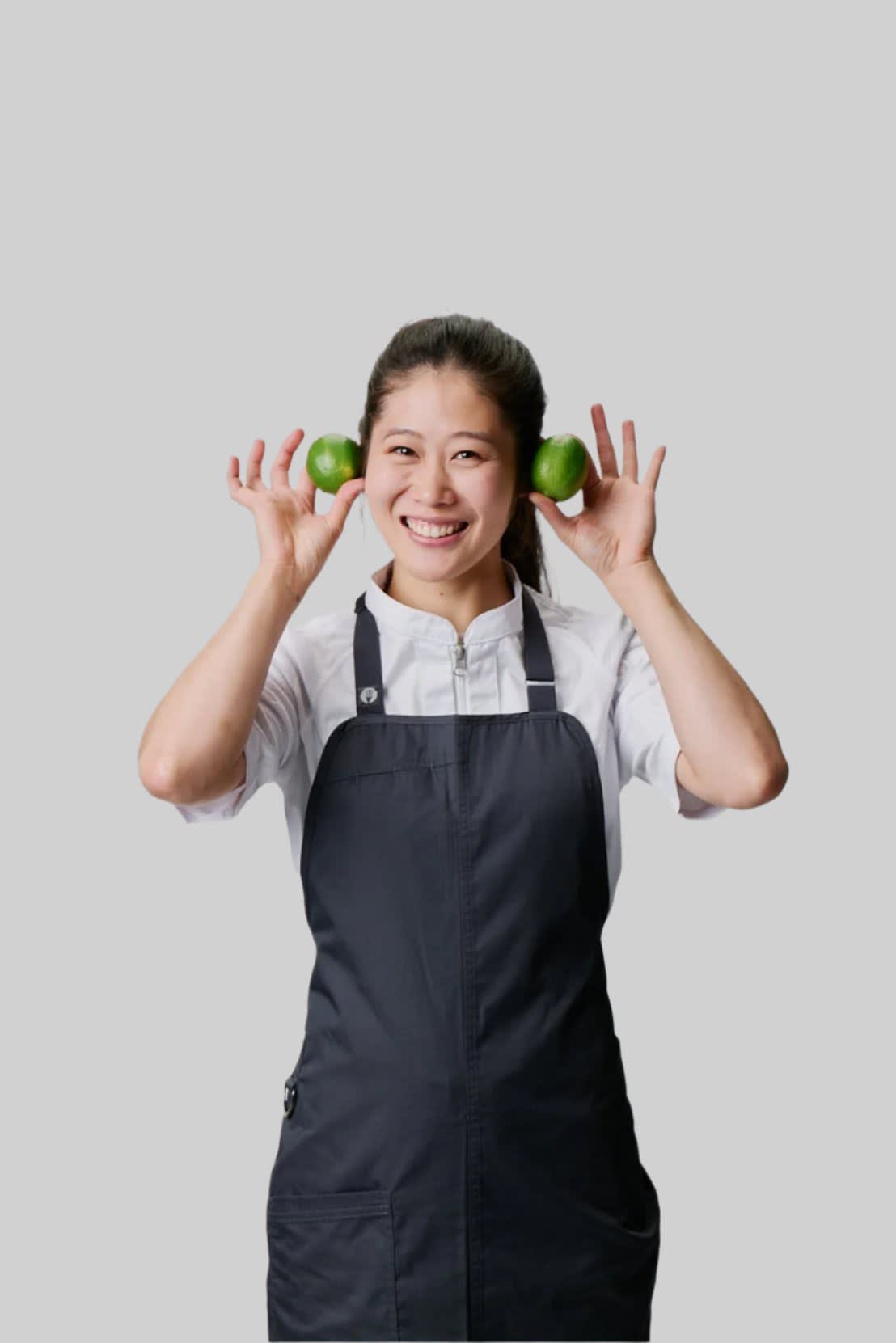 A woman in a black bib apron holds green limes to her ears, smiling against a neutral background.