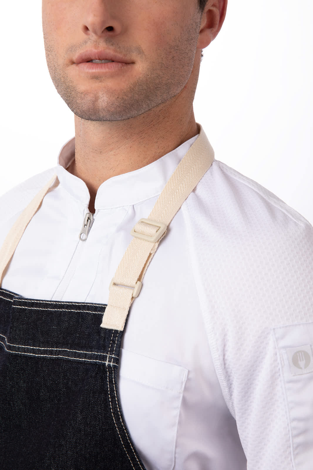 A denim bib apron with a beige adjustable strap is worn by a model. The fabric appears sturdy and durable.