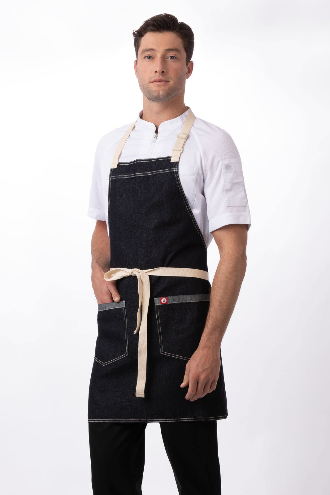 A man in a dark denim apron with cream straps, featuring pockets, stands against a white background.