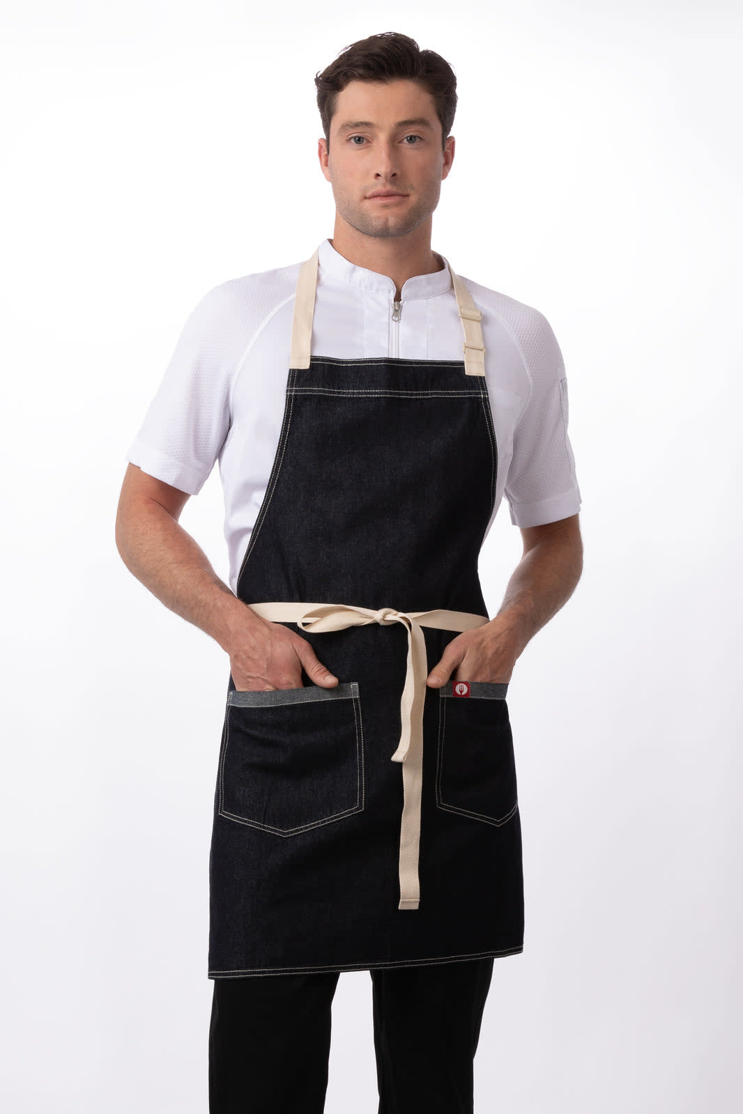 A denim bib apron in dark blue with a beige tie and pockets, worn by a model. Features visible stitching and a logo.