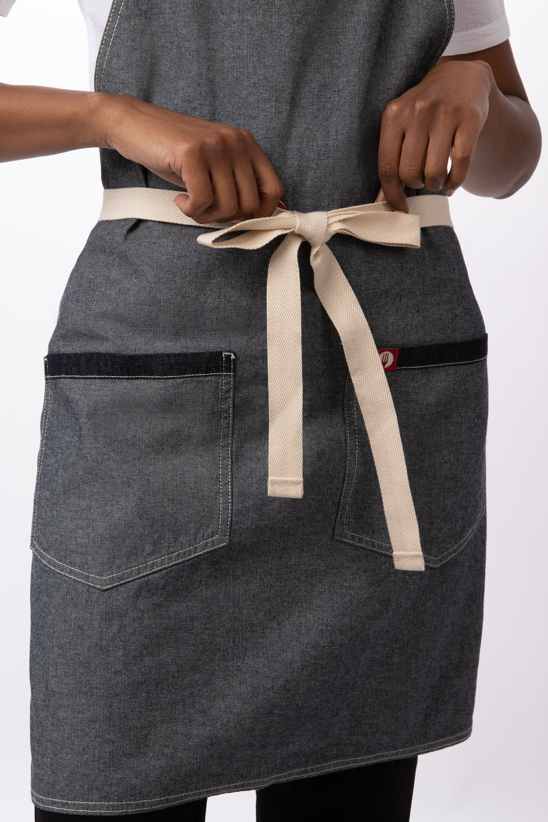 A dark denim bib apron with a light beige waist tie and pockets, featuring a branding logo.