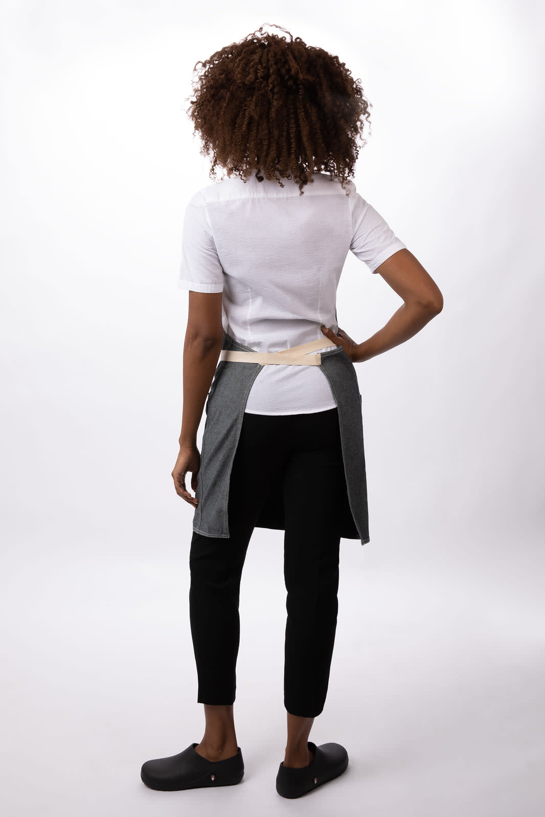 A person wearing a grey Ridgewood Bib Apron over a white shirt and black pants, with curly hair facing away from the camera.