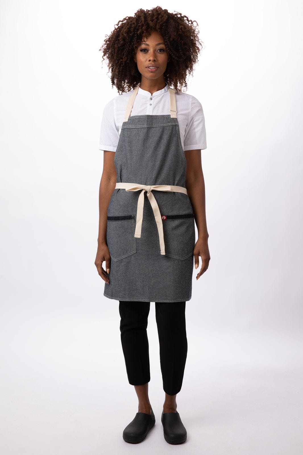 A grey bib apron with a cream tie and pockets, worn by a model in a white shirt and black trousers.
