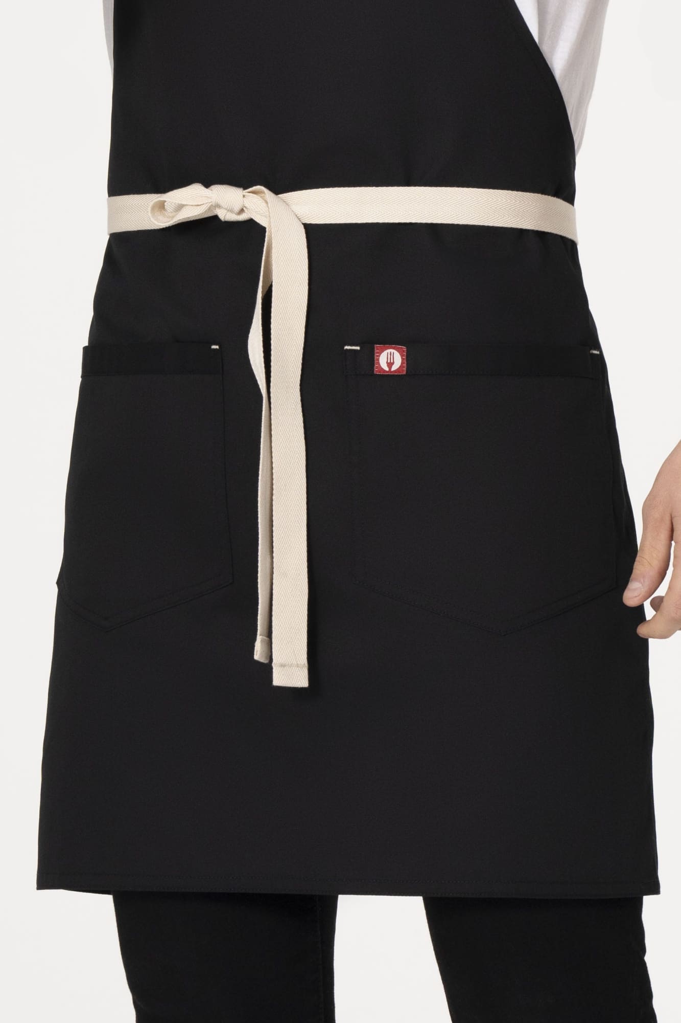 A black bib apron with a cream-coloured tie belt and pockets, featuring a small logo.