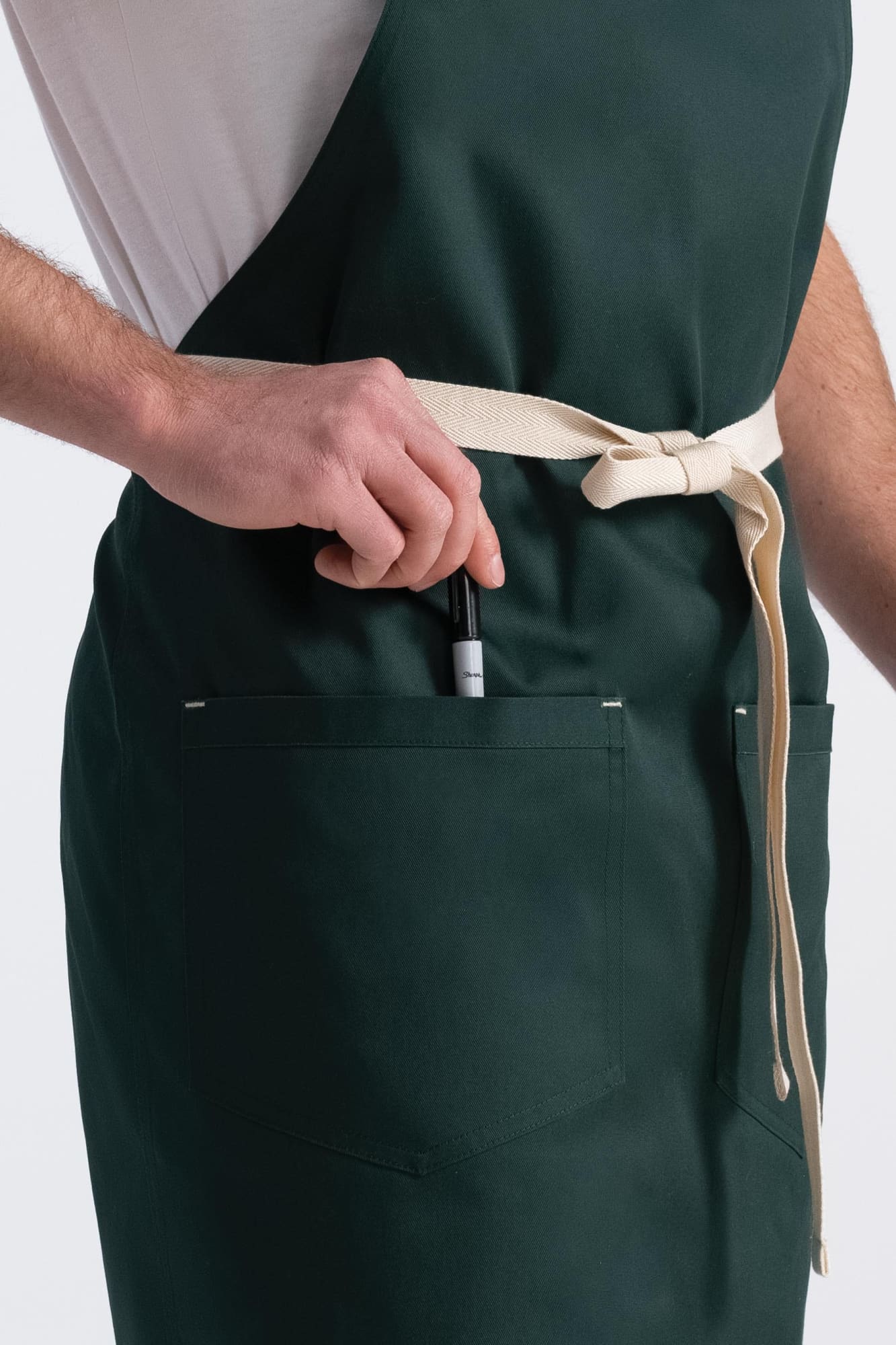 A dark green bib apron with a beige tie, featuring pockets, held by a person with a pen in one pocket.
