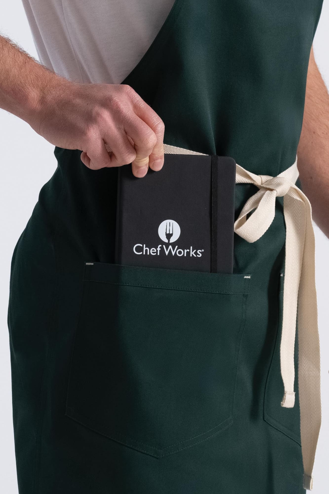 A forest green bib apron with a light-coloured tie, featuring a pocket holding a notebook with a logo.