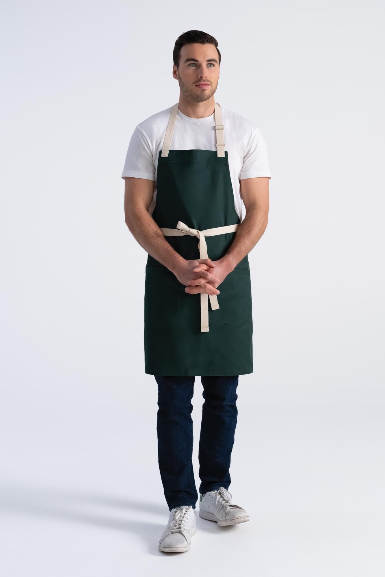 A green bib apron with beige straps worn over a white T-shirt, modelled by a standing man.