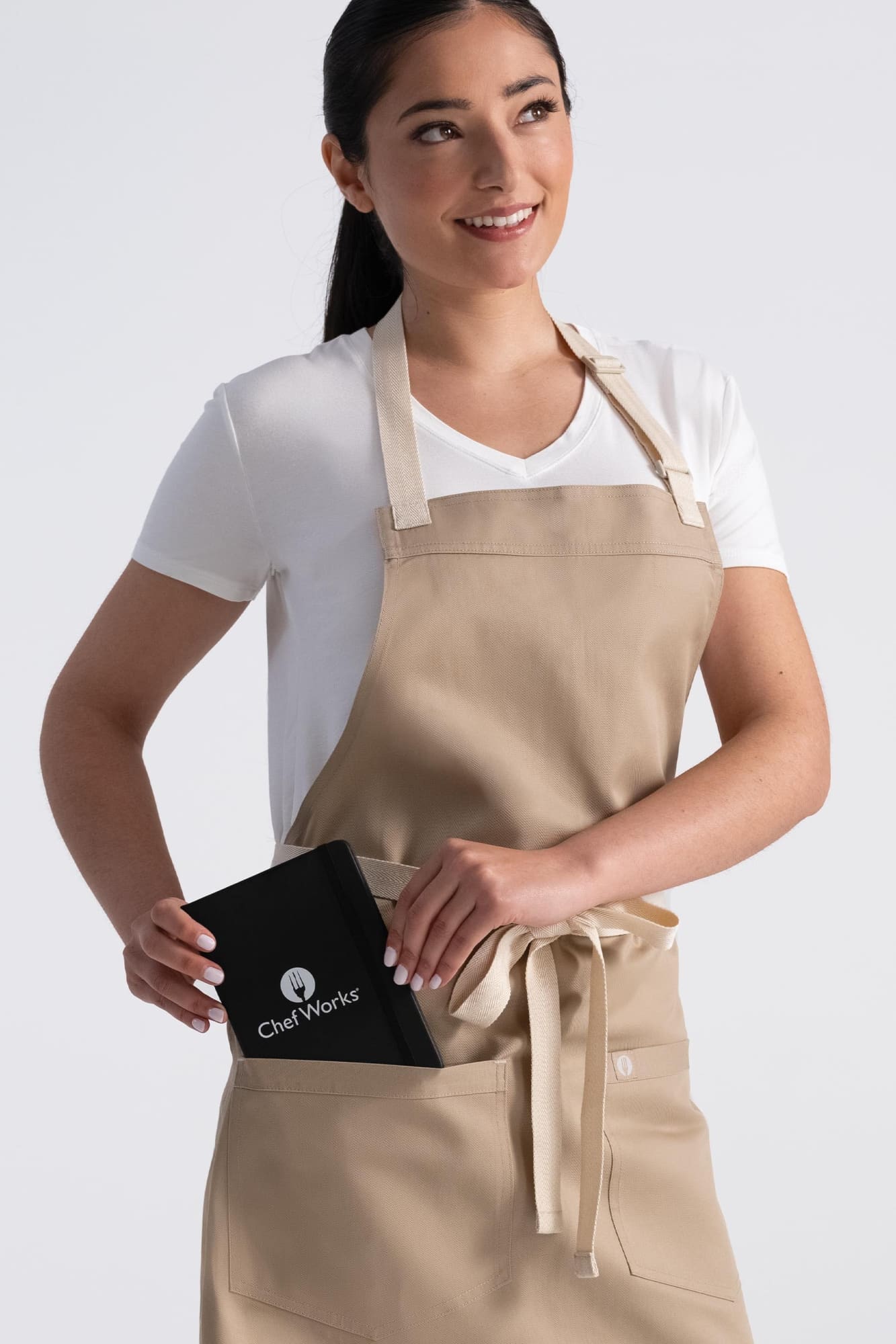 A woman wearing a beige Ridgewood Bib Apron holds a black item in the front pocket, with a logo visible.
