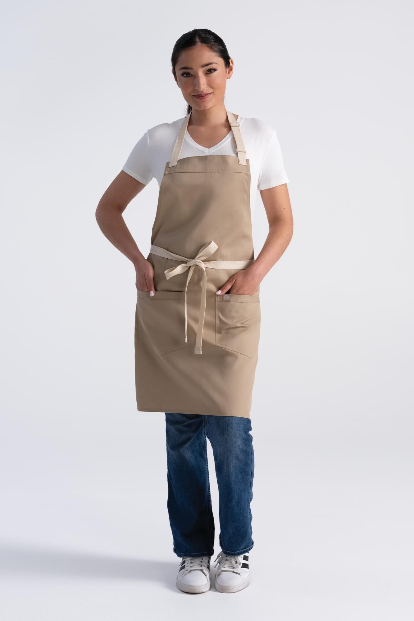 A woman wearing a beige bib apron with pockets, standing with hands in her pockets, against a plain background.