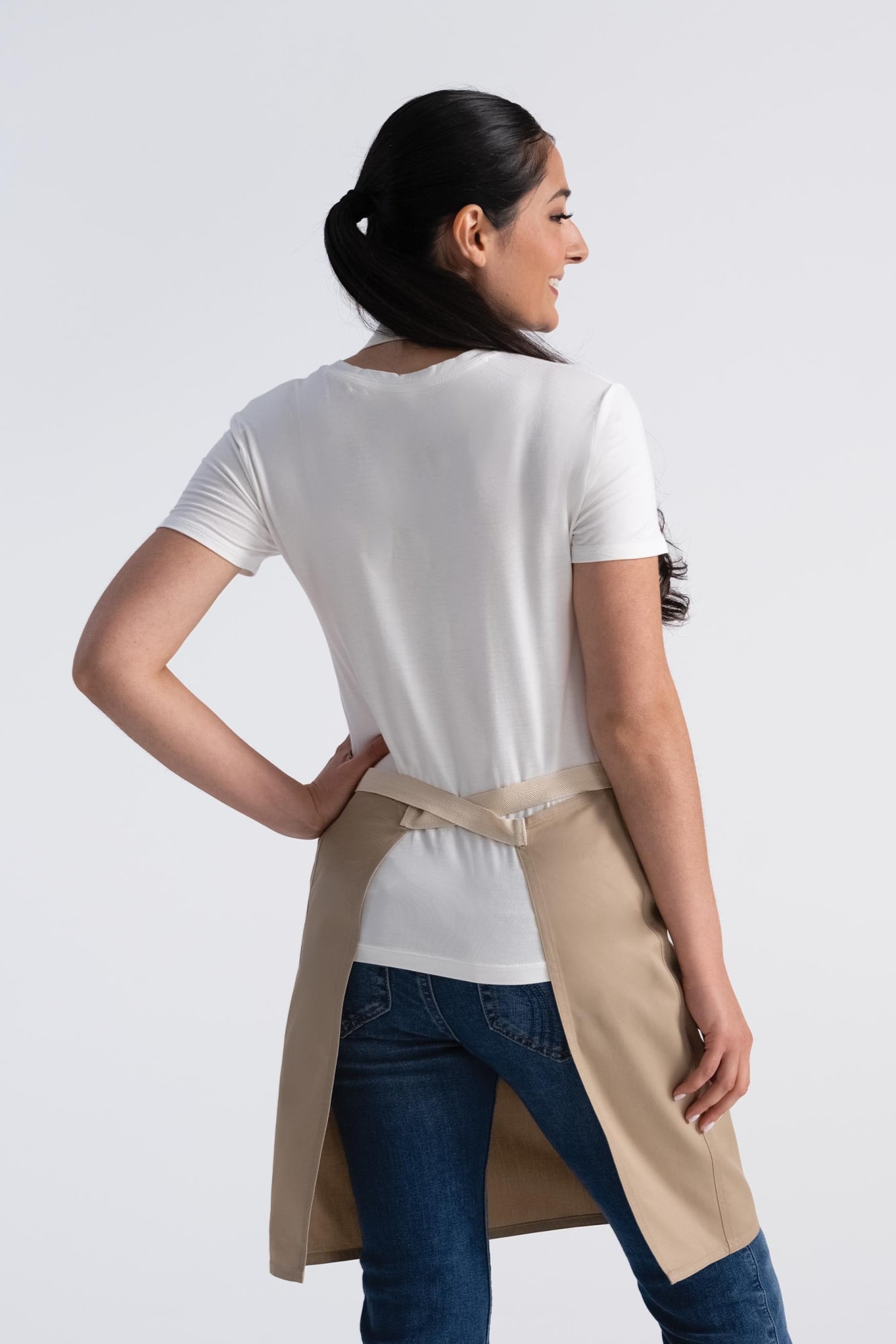 A woman wearing a beige Ridgewood Bib Apron over a white t-shirt, facing away from the camera.