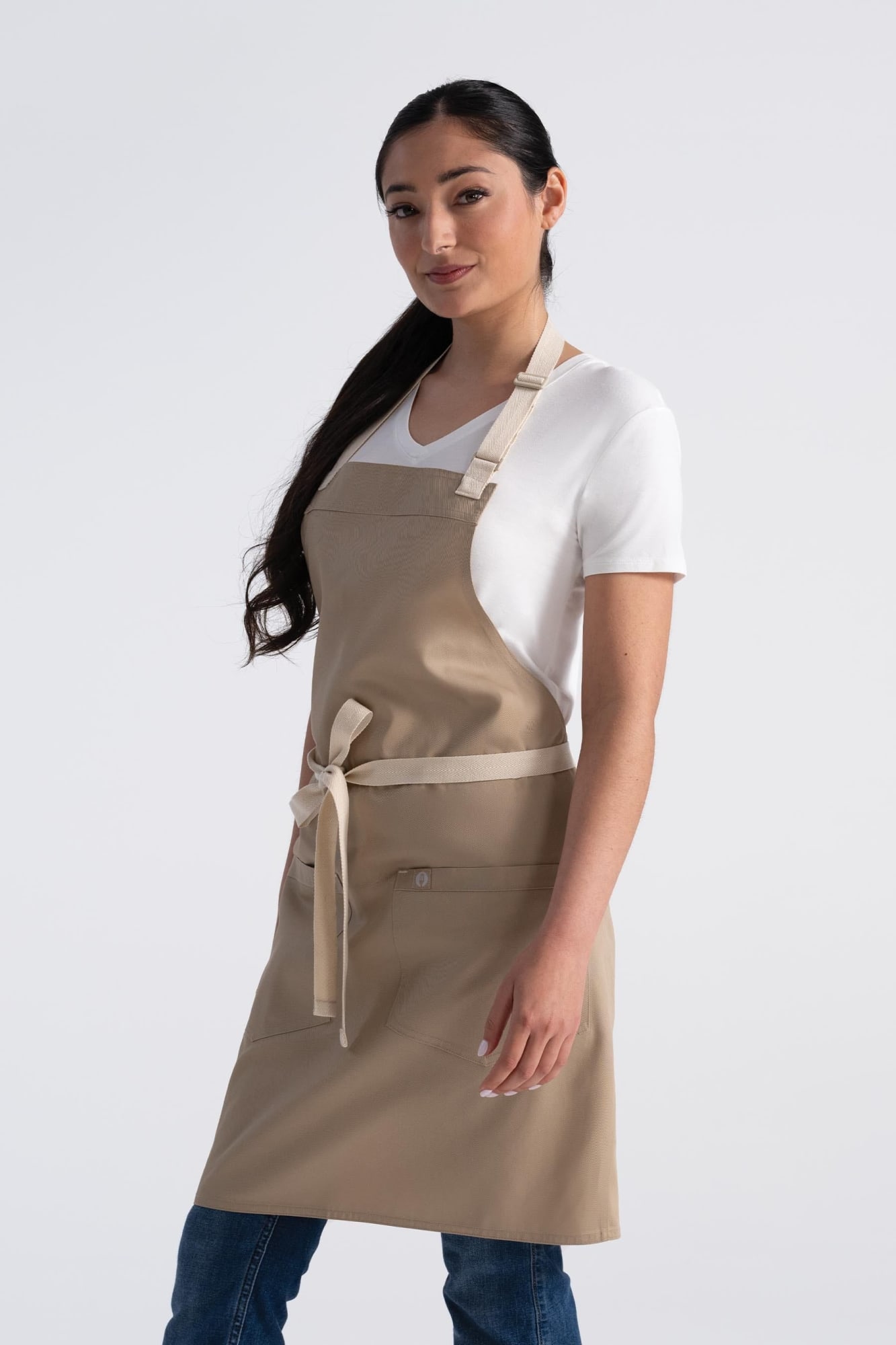 A woman wearing a beige Ridgewood Bib Apron, featuring a front pocket and a tie at the waist.