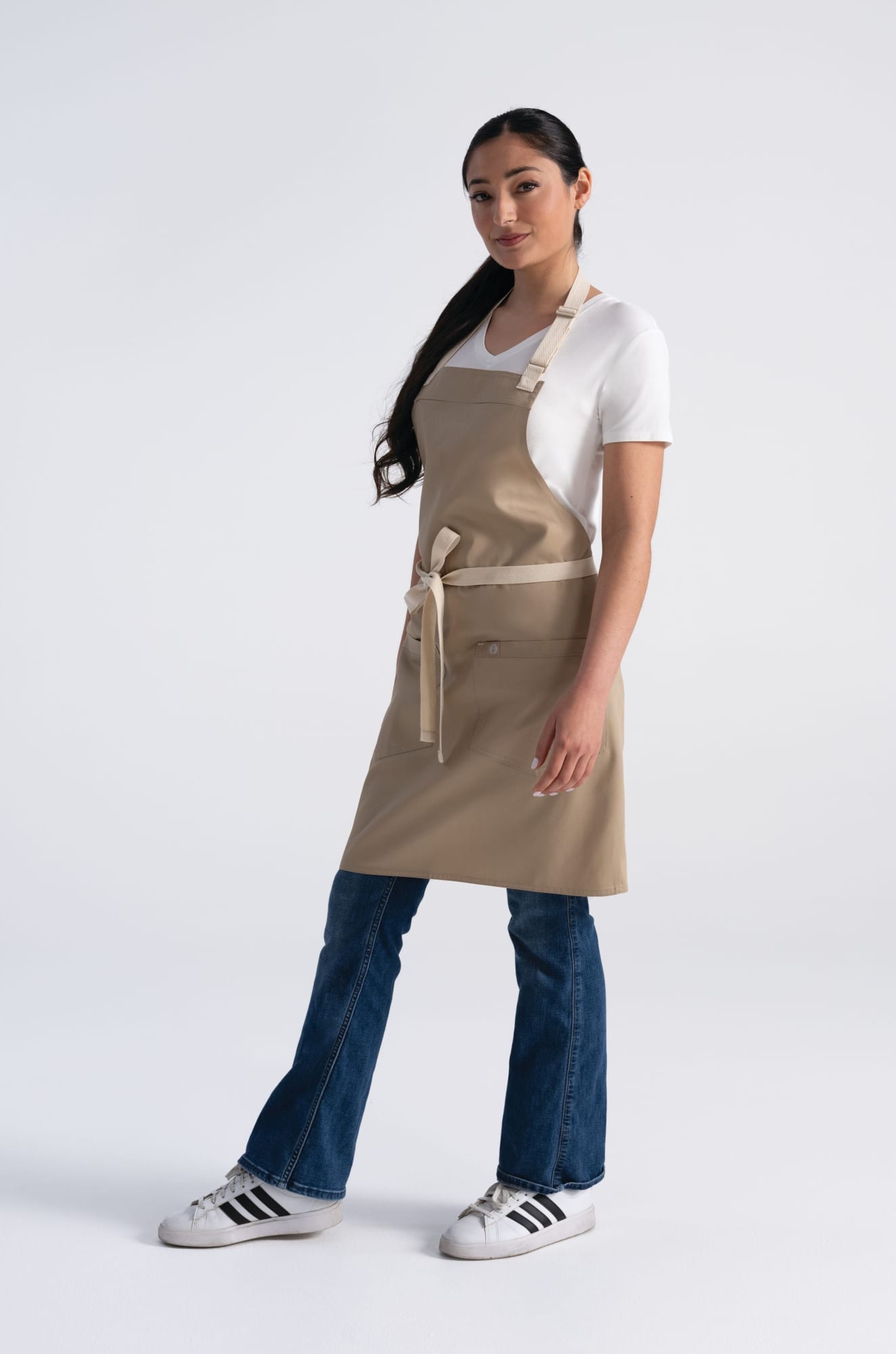 A beige bib apron is worn by a model, who pairs it with a white top and blue jeans. The apron features a logo.