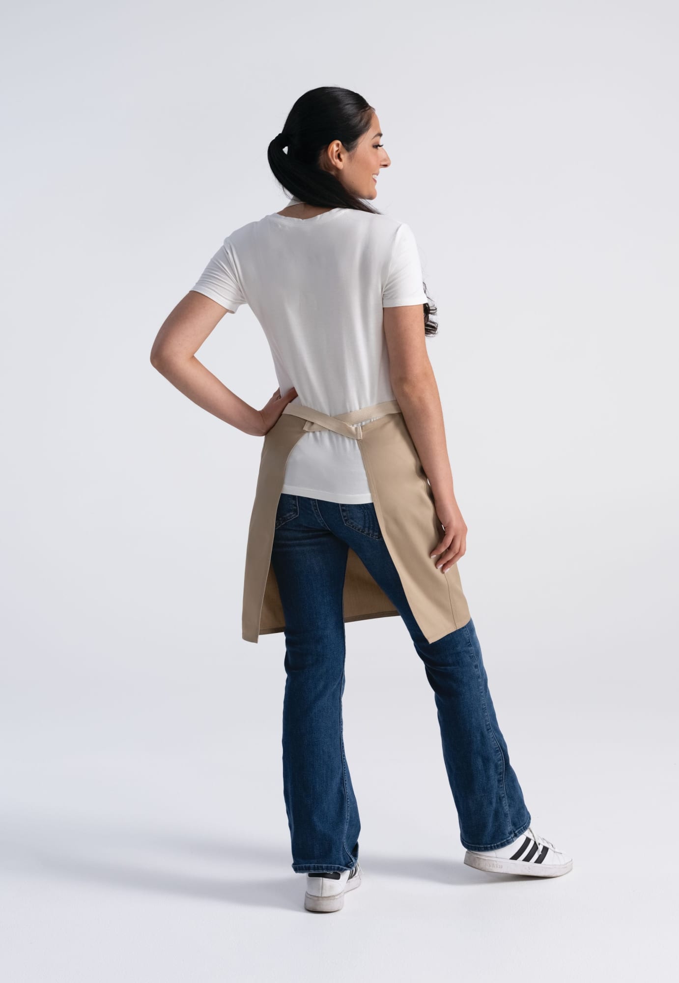 A woman wearing a beige Ridgewood Bib Apron over a white t-shirt, standing with her back to the camera, styled with blue jeans.