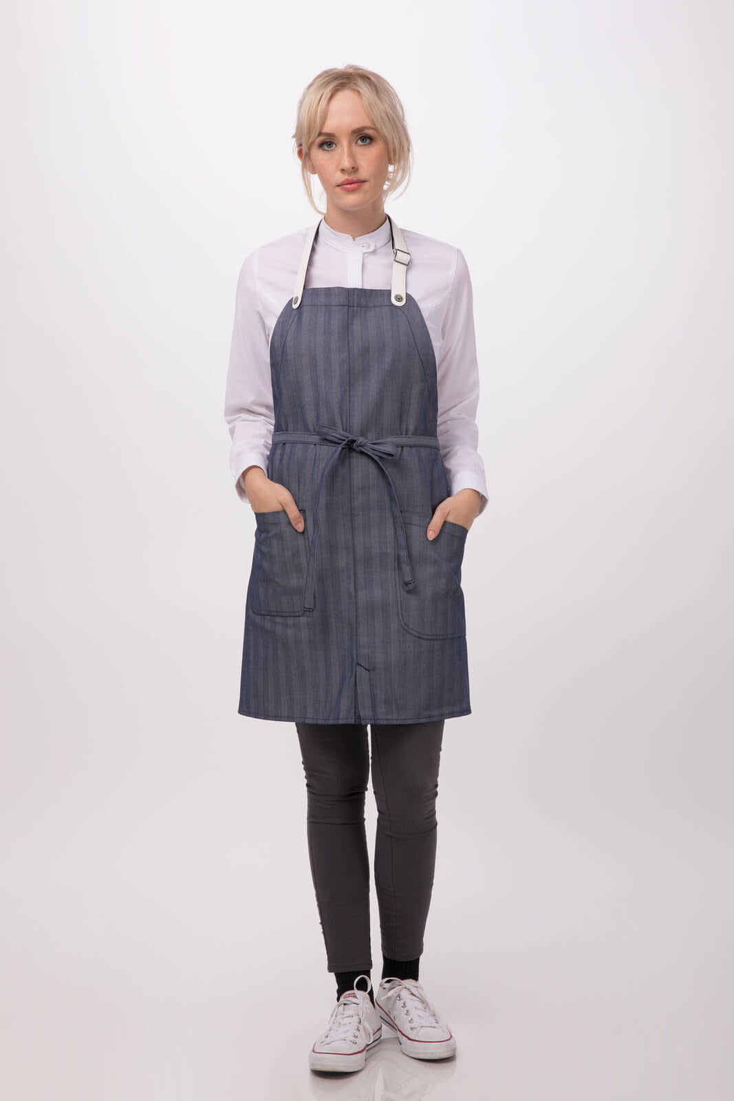 A woman wearing a navy blue bib apron with stripes, pockets, and a tie waist, over a white shirt and black pants.