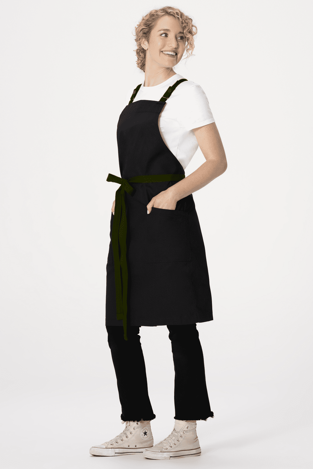 A black cross back apron with pockets worn over a white shirt, modelled by a person standing in profile.
