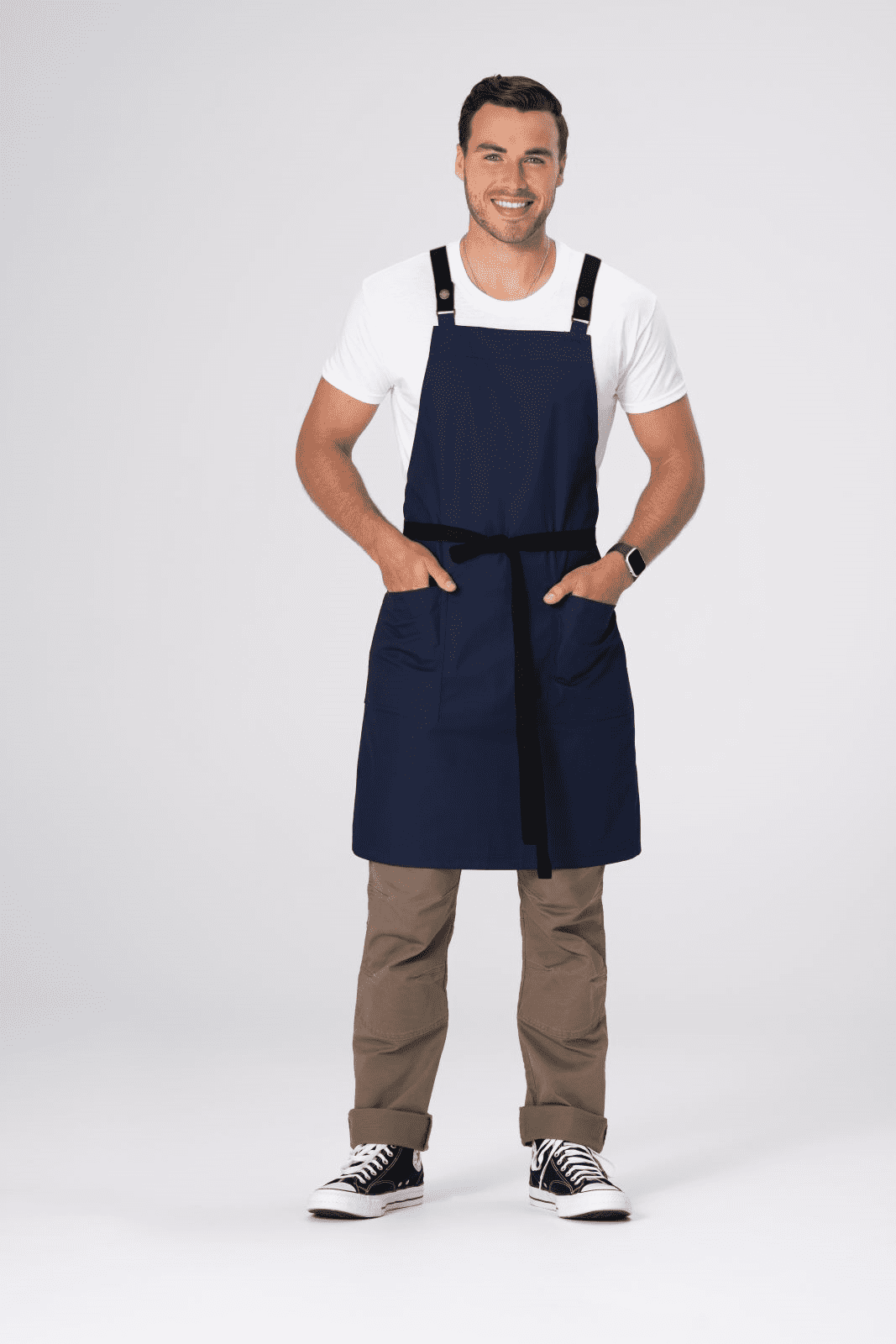 A navy blue cross back apron featuring adjustable straps and front pockets, modelled by a smiling man.