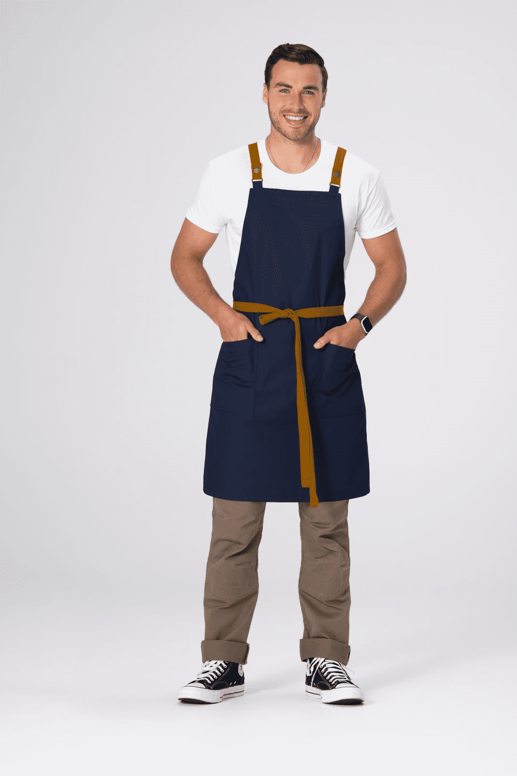 A cross back apron in navy with tan straps and a waist tie, featuring a model standing with hands in pockets.