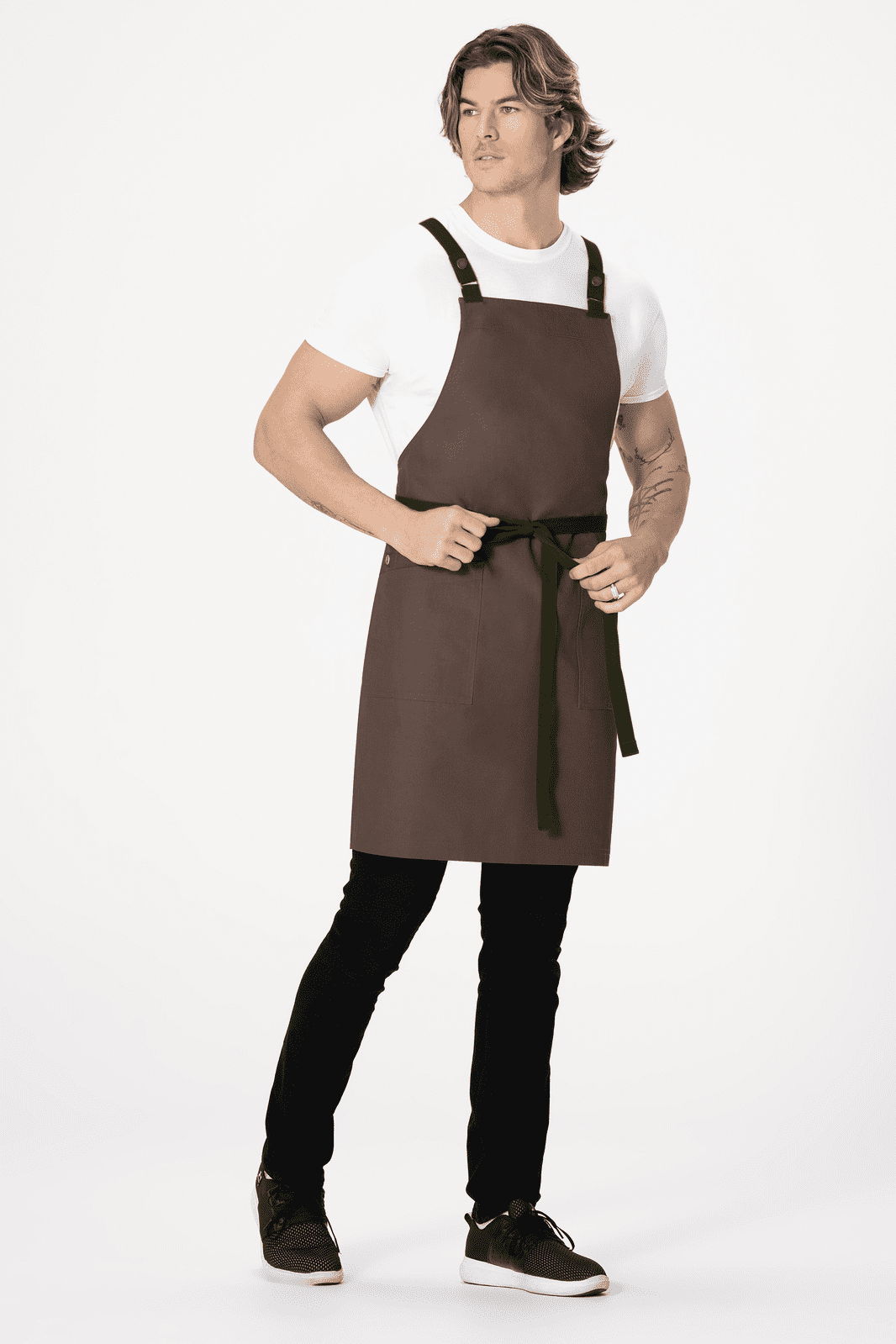 A brown cross back apron with a waist tie, worn by a model in a white t-shirt and black pants, featuring a logo.