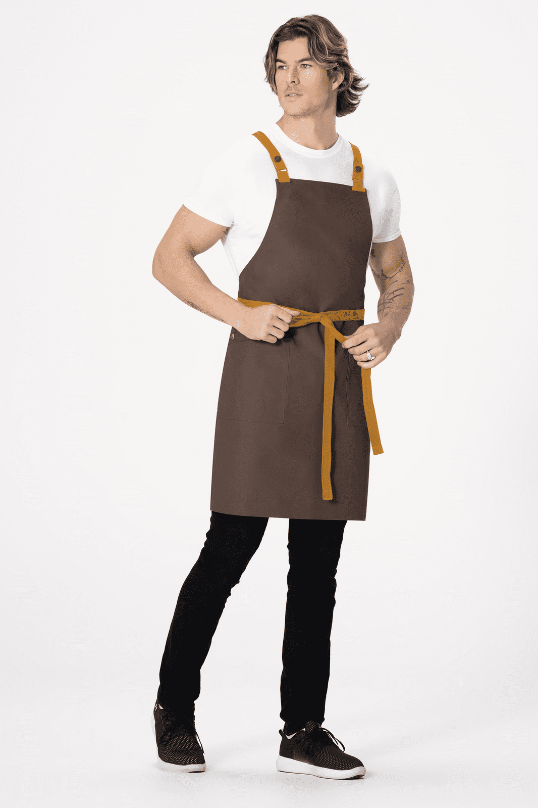 A brown cross-back apron with yellow straps, worn by a model standing in a neutral setting.