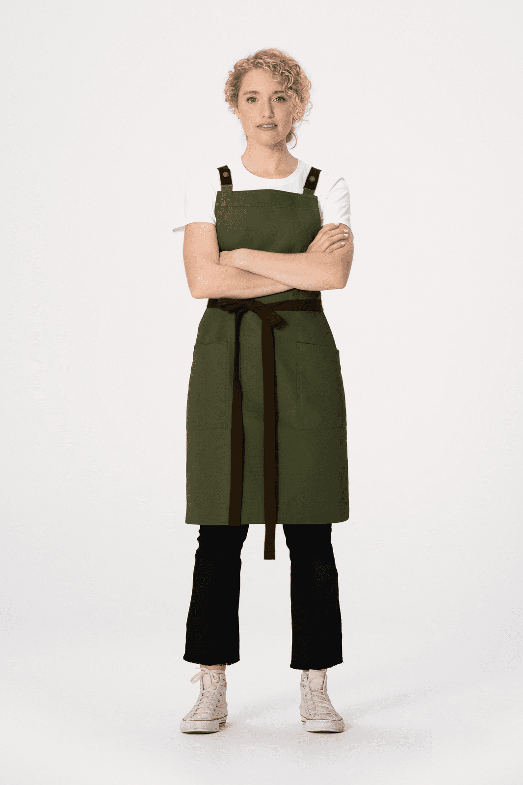 A woman in a green Lockharte Cross Back Apron stands with arms crossed, wearing a white shirt and black pants.