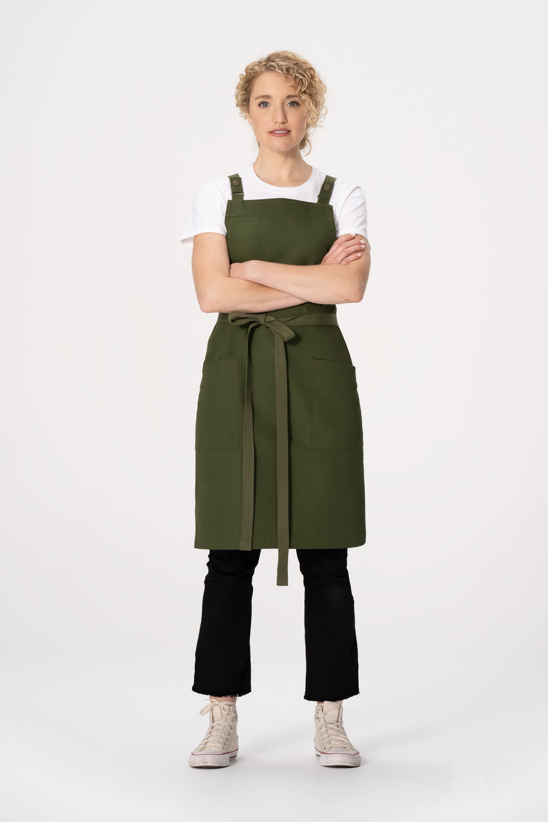 A green cross back apron with a tie waist, worn over a white t-shirt, modelled by a standing woman with curly hair.