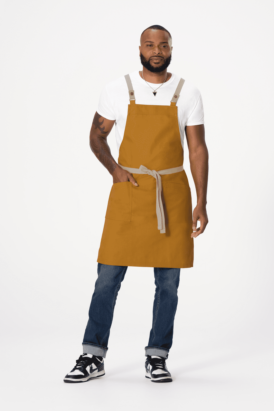 A man wearing a mustard-coloured cross back apron with a grey tie, denim jeans, and sneakers. The apron has a logo.
