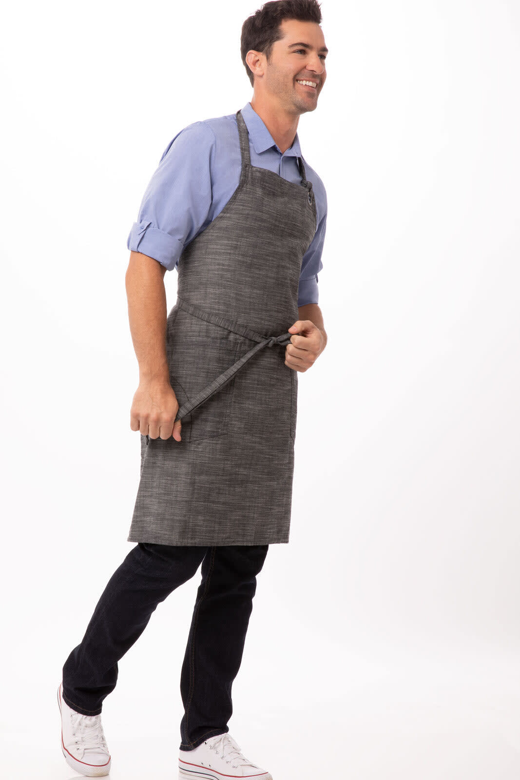 A man in a grey Corvallis bib apron stands confidently, holding a utensil, wearing a light blue shirt and dark jeans.
