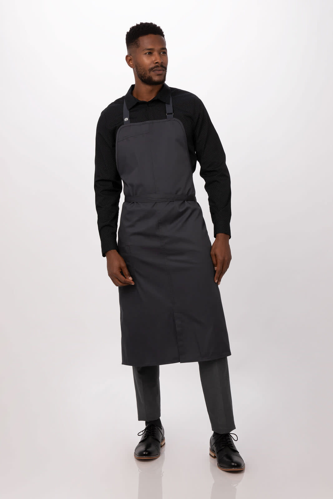 A grey bib apron worn by a man, styled with a black shirt and matching trousers. The design includes straps and pockets.
