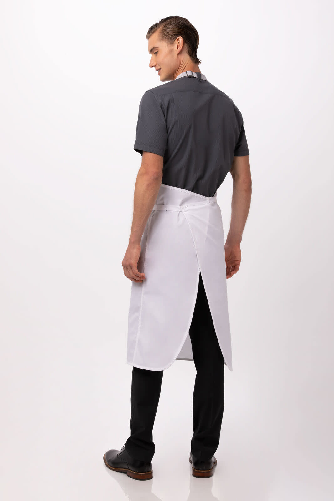 A man wearing a white bib apron over a grey shirt, viewed from the back with black trousers and shoes.