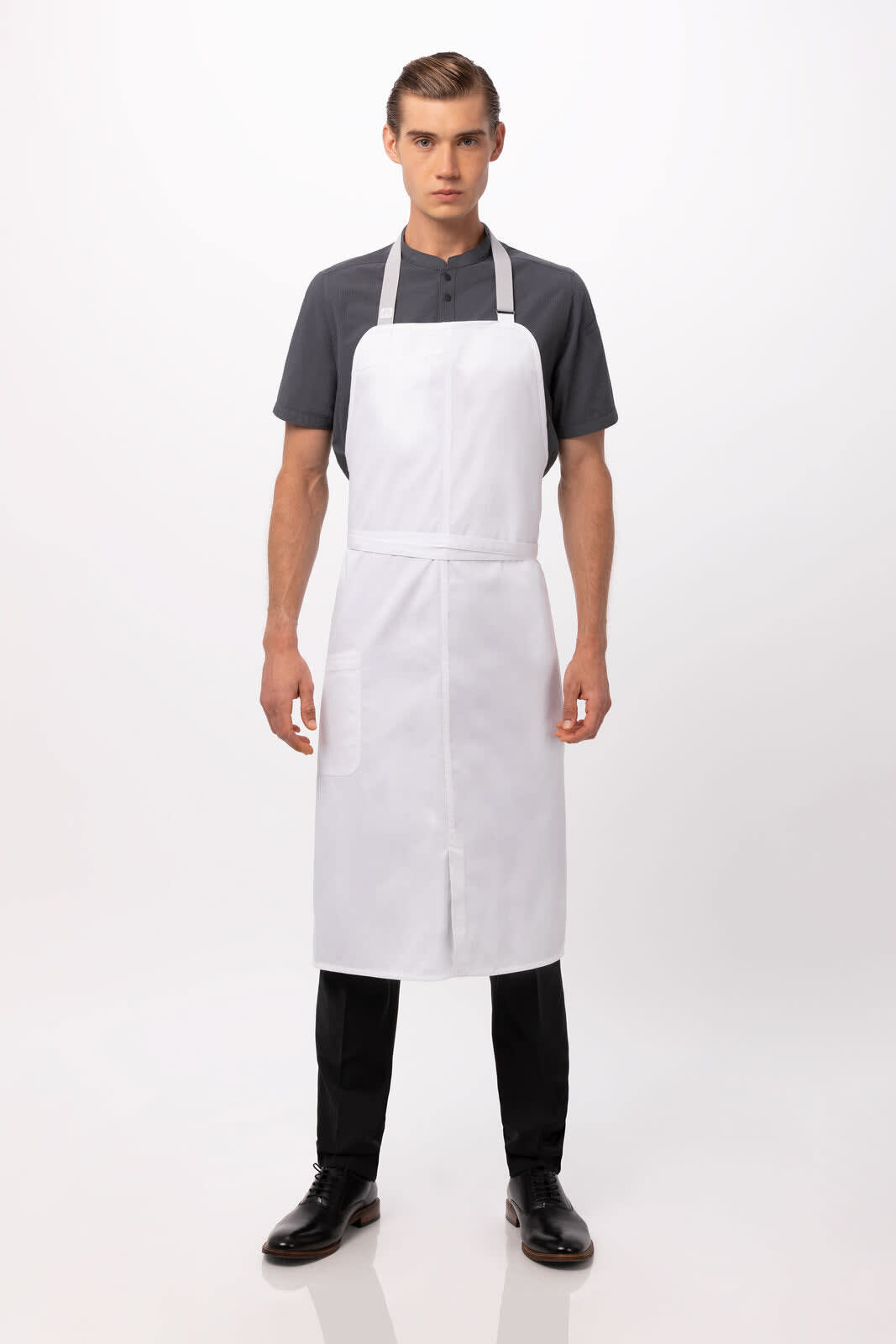 A white bib apron worn by a model, featuring a simple design and adjustable straps. The model is dressed in black trousers and shoes.