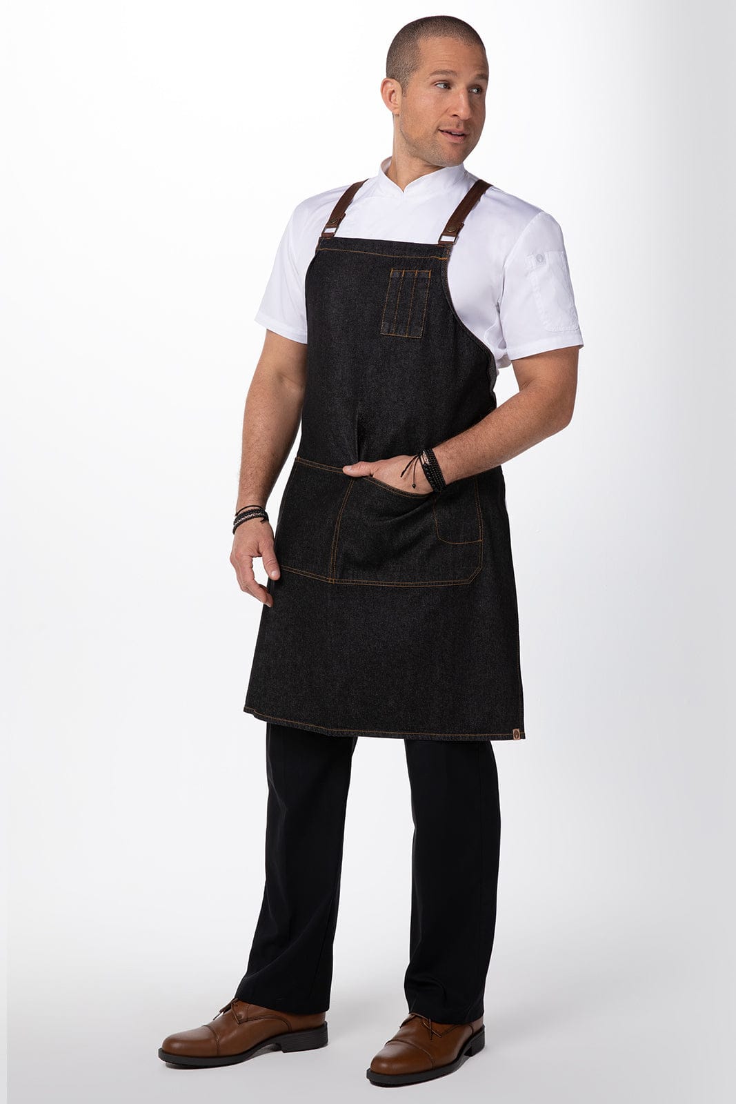 A man stands wearing a black denim cross back apron over a white shirt and black trousers, featuring pockets and a branding logo.