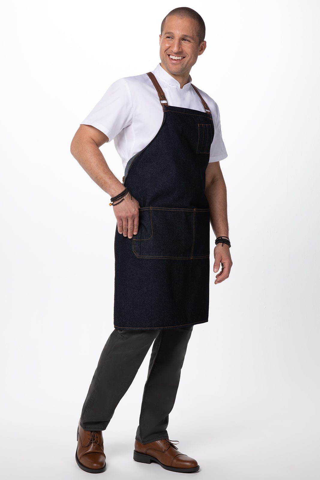 A denim cross-back apron in dark blue, featuring pockets and leather straps, worn by a smiling man.