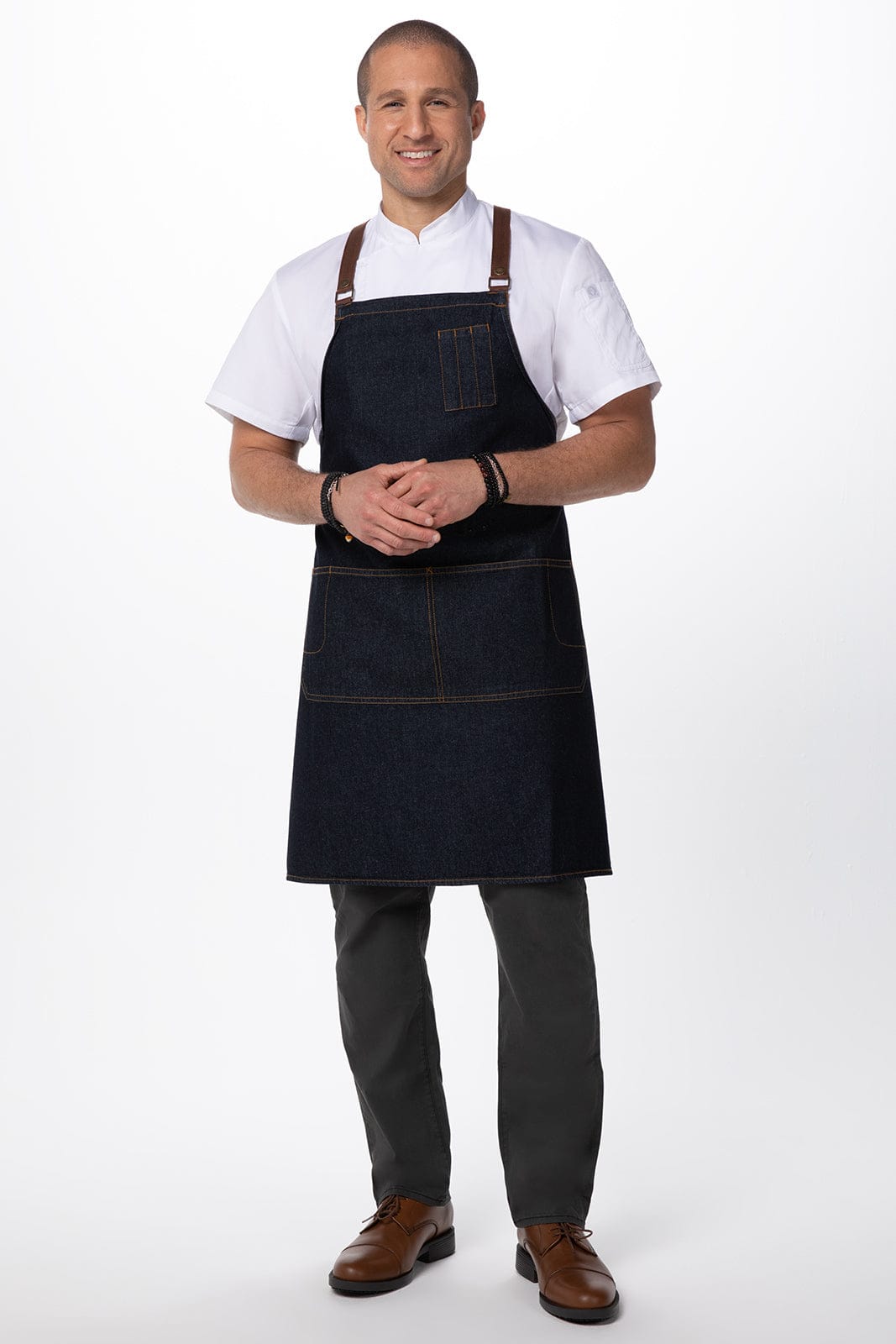 A man wearing a dark denim cross back apron, featuring pockets and brown straps, over a white shirt and grey pants.