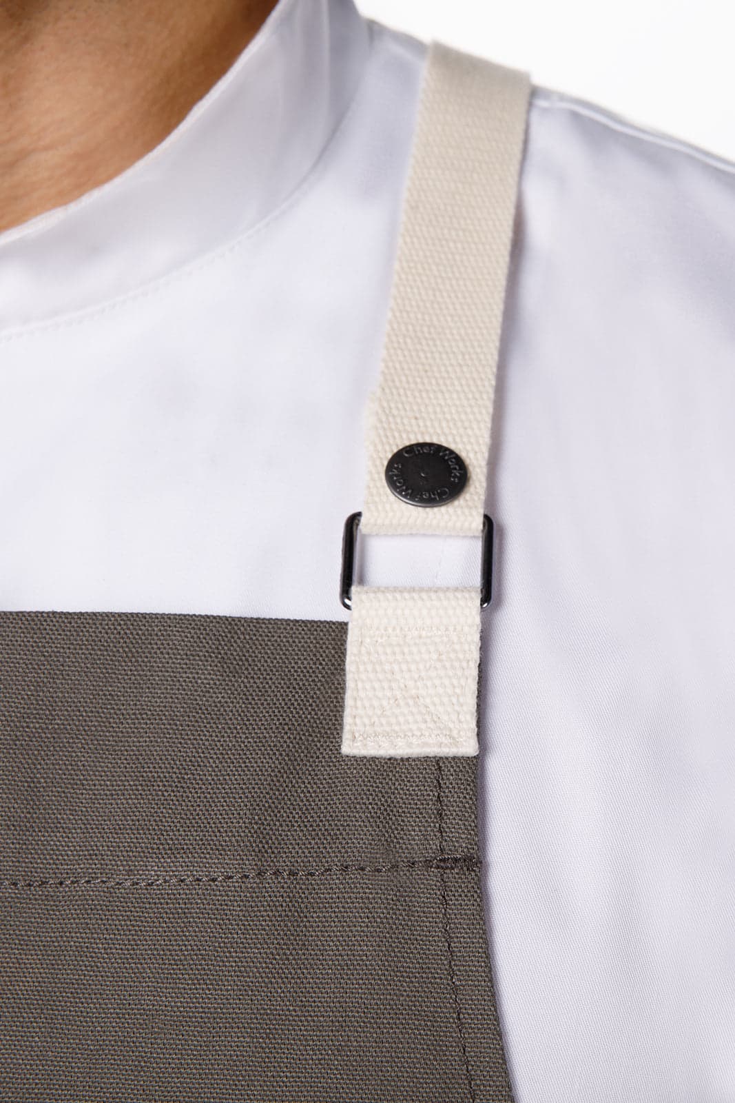 A grey Byron Cross-Back Apron featuring a cream-coloured strap and a black fastener. Includes a branding logo.