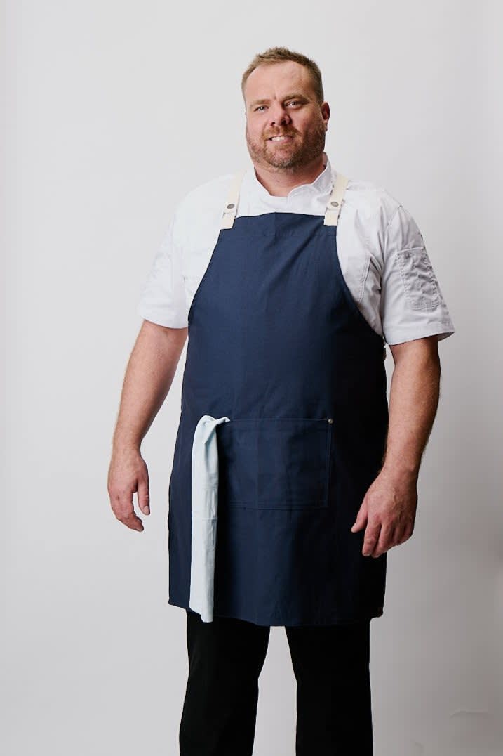 A navy cross-back apron displayed on a model wearing a white shirt, featuring a logo and a front pocket.