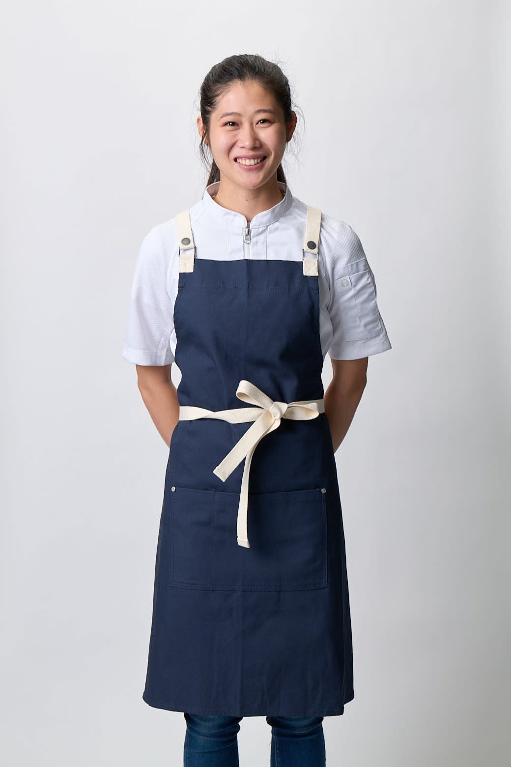 A navy blue apron with a cream-coloured tie, featuring adjustable straps and pockets, displayed on a model.
