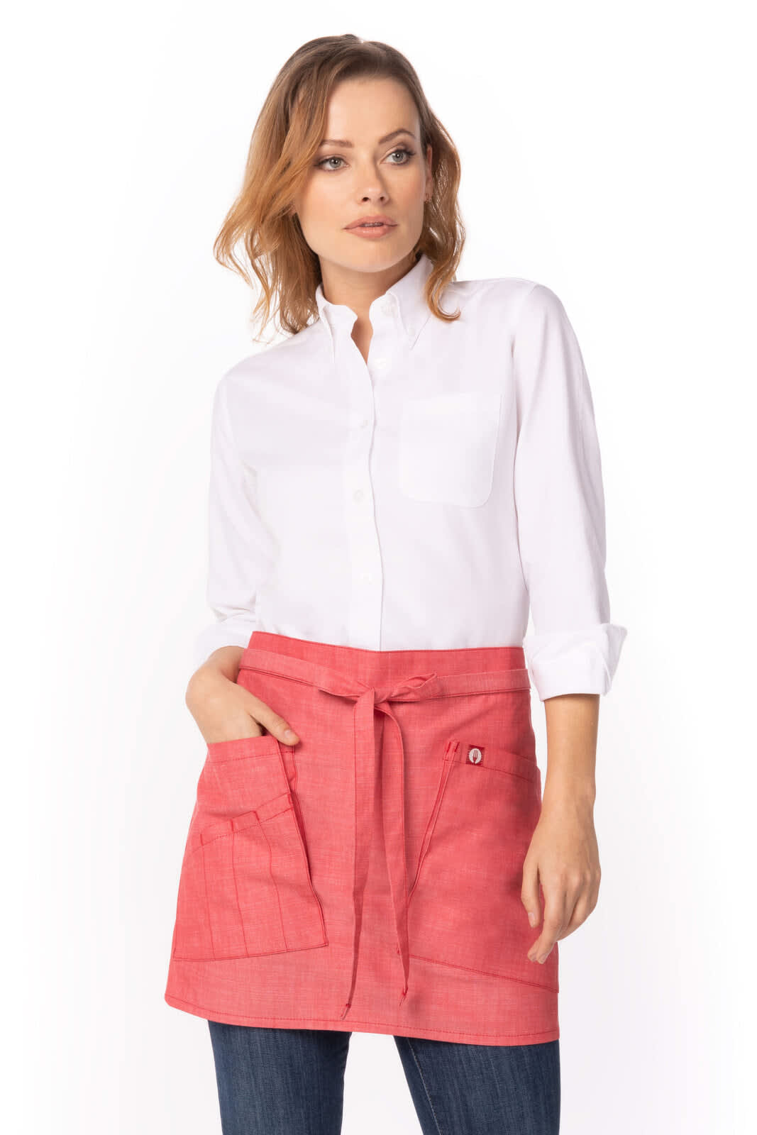 A woman wearing a coral bistro apron over a white shirt, featuring pockets and a waist tie. The apron has a logo.