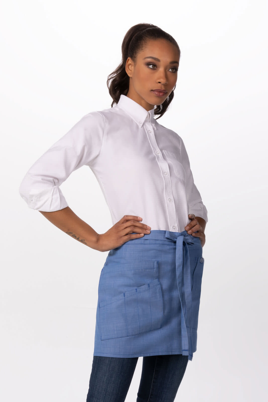 A woman wears a light blue half bistro apron over a white shirt and jeans, featuring pockets and a tied waist.