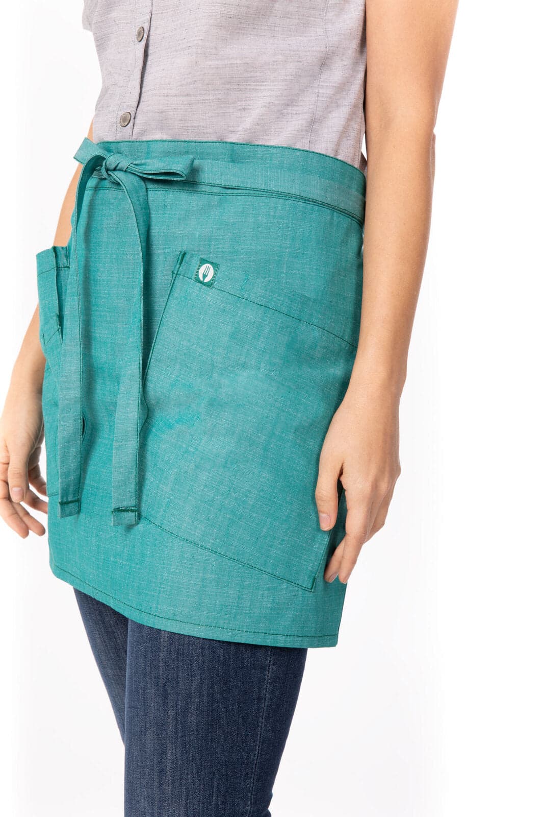 A teal bistro apron features a waist tie and front pocket, shown on a model with denim jeans.