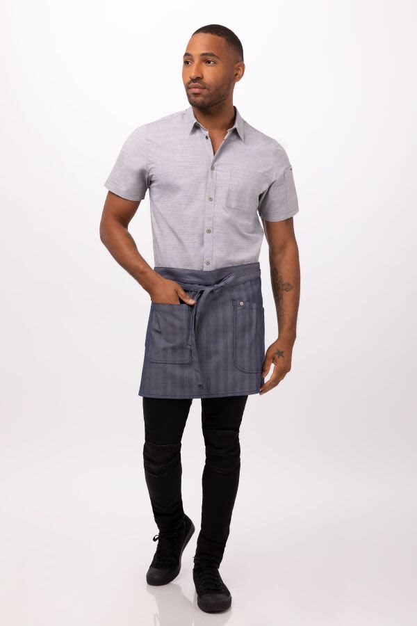 A man wears a grey short-sleeve shirt and a navy striped half bistro apron, standing in a neutral setting.
