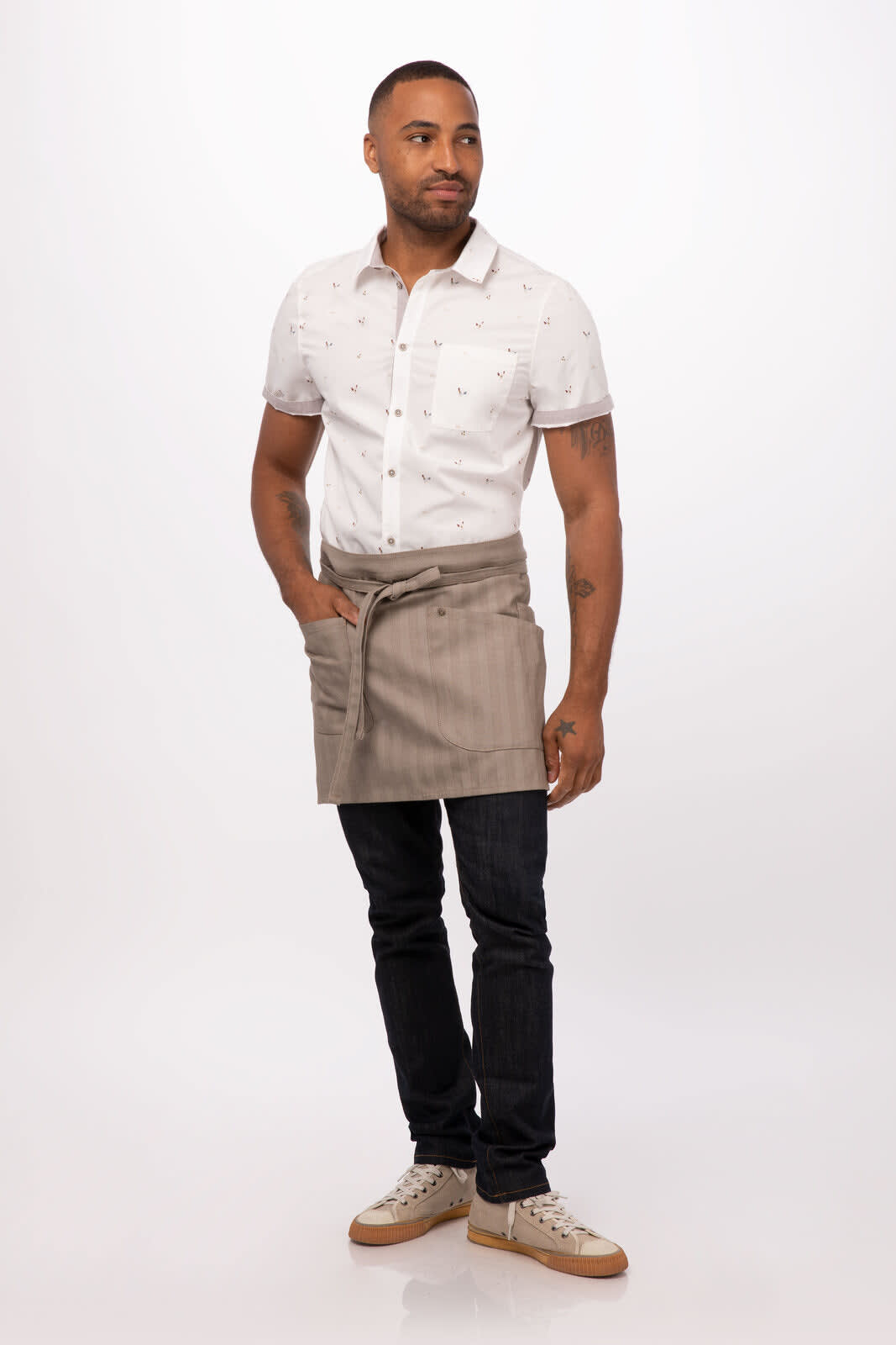 A model wearing a grey striped half bistro apron over a white patterned shirt, paired with dark jeans and sneakers.
