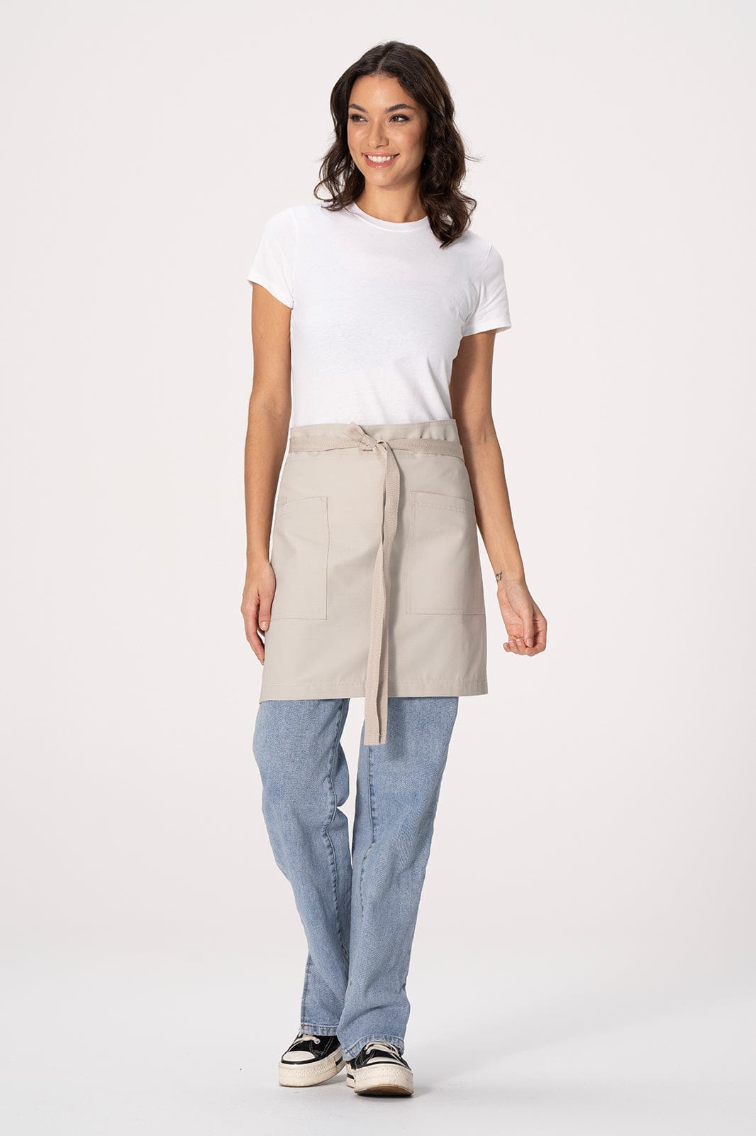 A waist apron in light beige, featuring a tied waistband, worn over blue jeans by a model. The apron includes a logo.