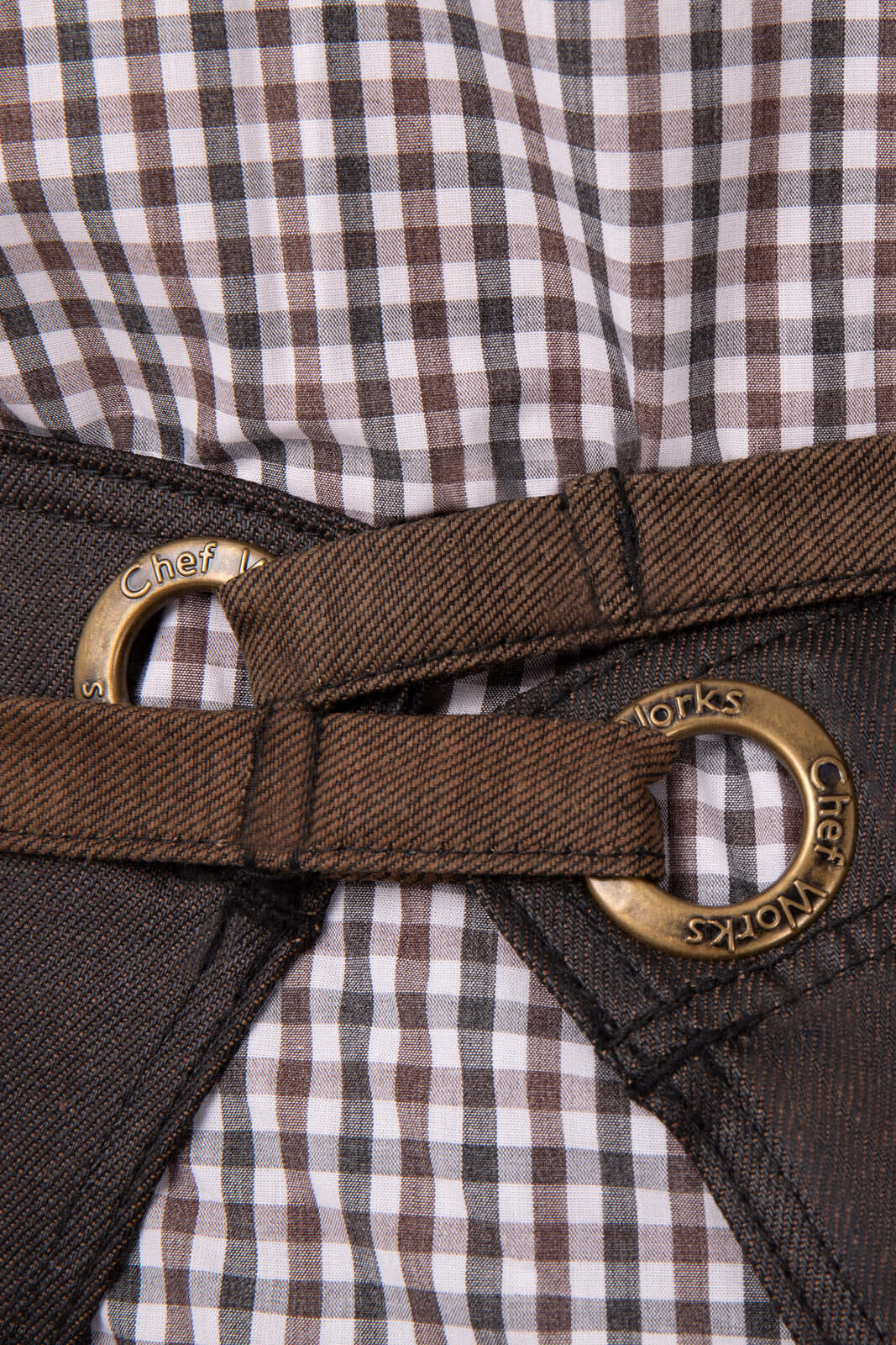 A brown apron features metal eyelets and a checkered fabric underneath, showcasing its stylish design and sturdy construction.
