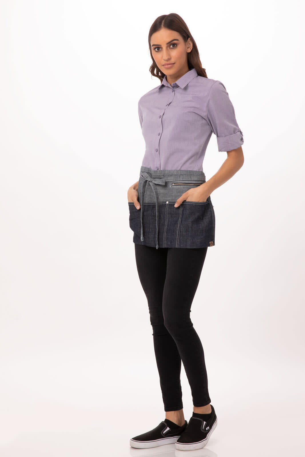 A person wearing a denim waist apron with pockets, styled with a light purple shirt and black pants.