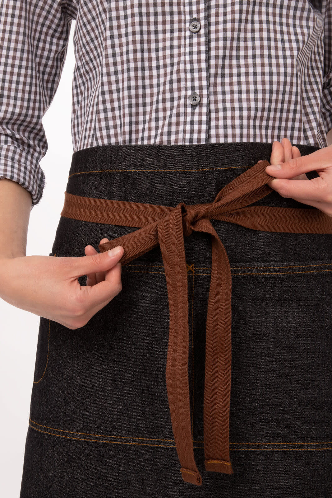 A half bistro apron in dark denim, featuring a brown waist tie and pockets, being tied by a person.